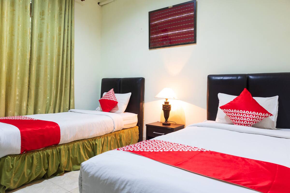 Une chambre d’hôtel avec deux lits, chacun recouvert d’une couverture rouge et blanche, une table de chevet avec une lampe et une fenêtre avec des rideaux.