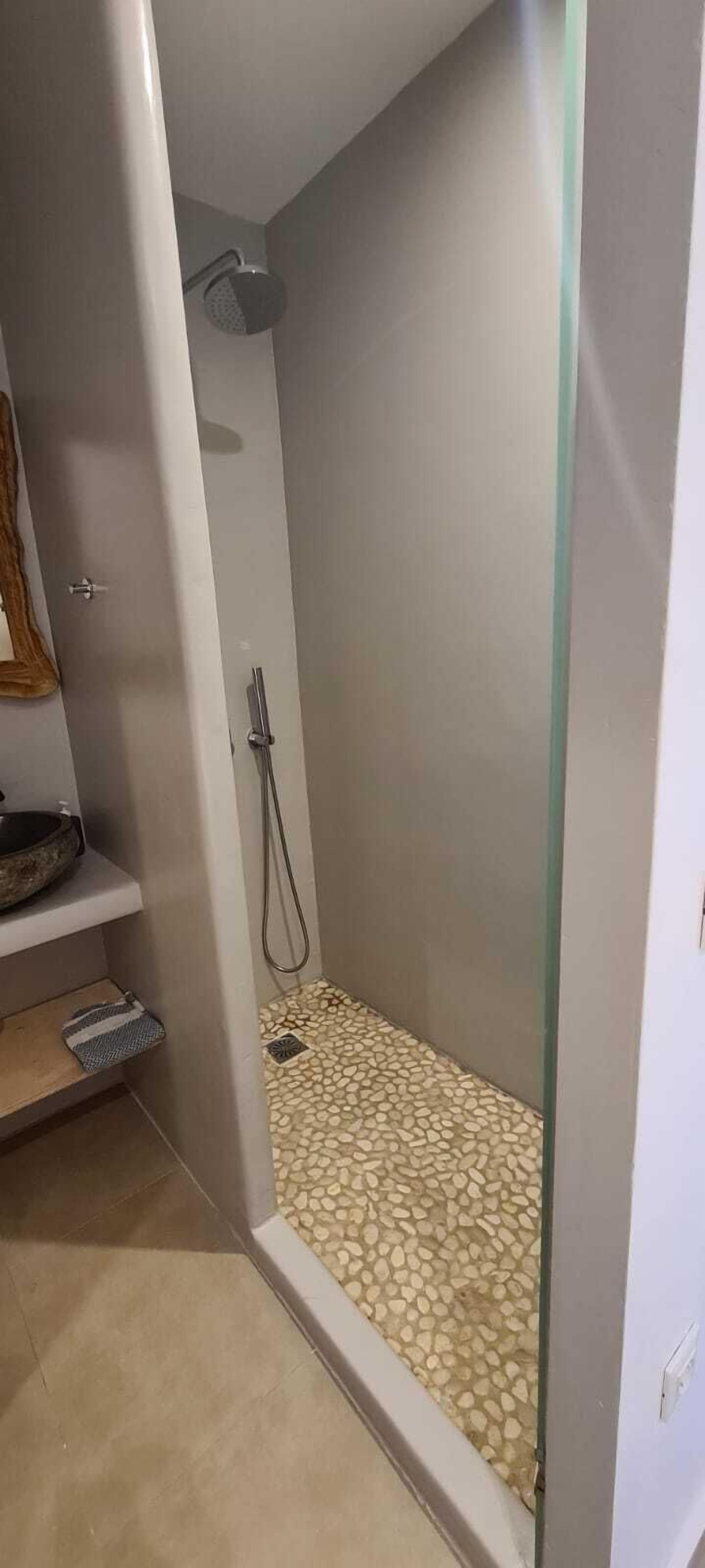Een kleine, afgesloten douche ruimte met een vloer van kiezelstenen, een handdouche en een klein plankje met een kom.
