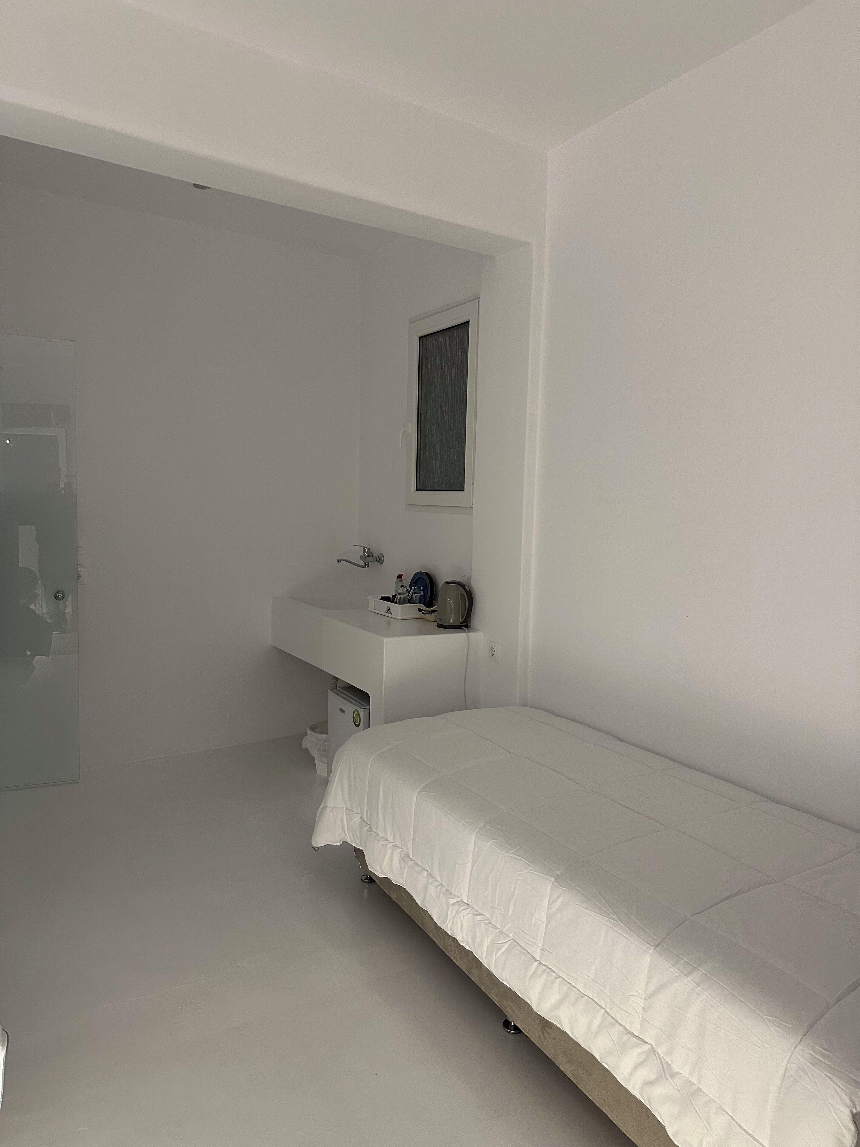 Una habitación pequeña y blanca con una cama individual, una mesita de noche, una ventana y una puerta.