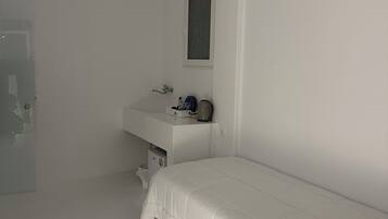 Una habitación pequeña y blanca con una cama individual, una mesita de noche, una ventana y una puerta.
