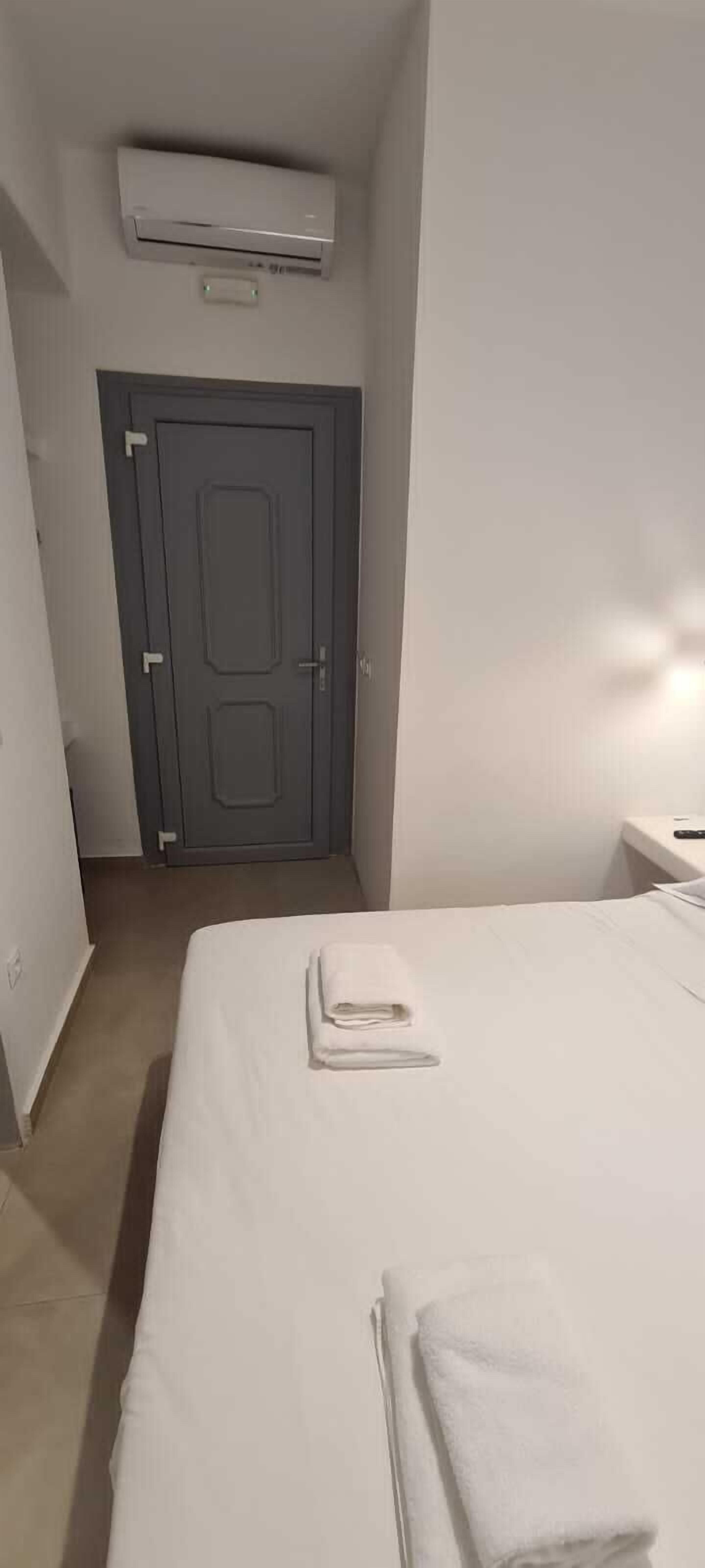 A hotel room with a bed, a bedside table, an air conditioning unit, and a door.