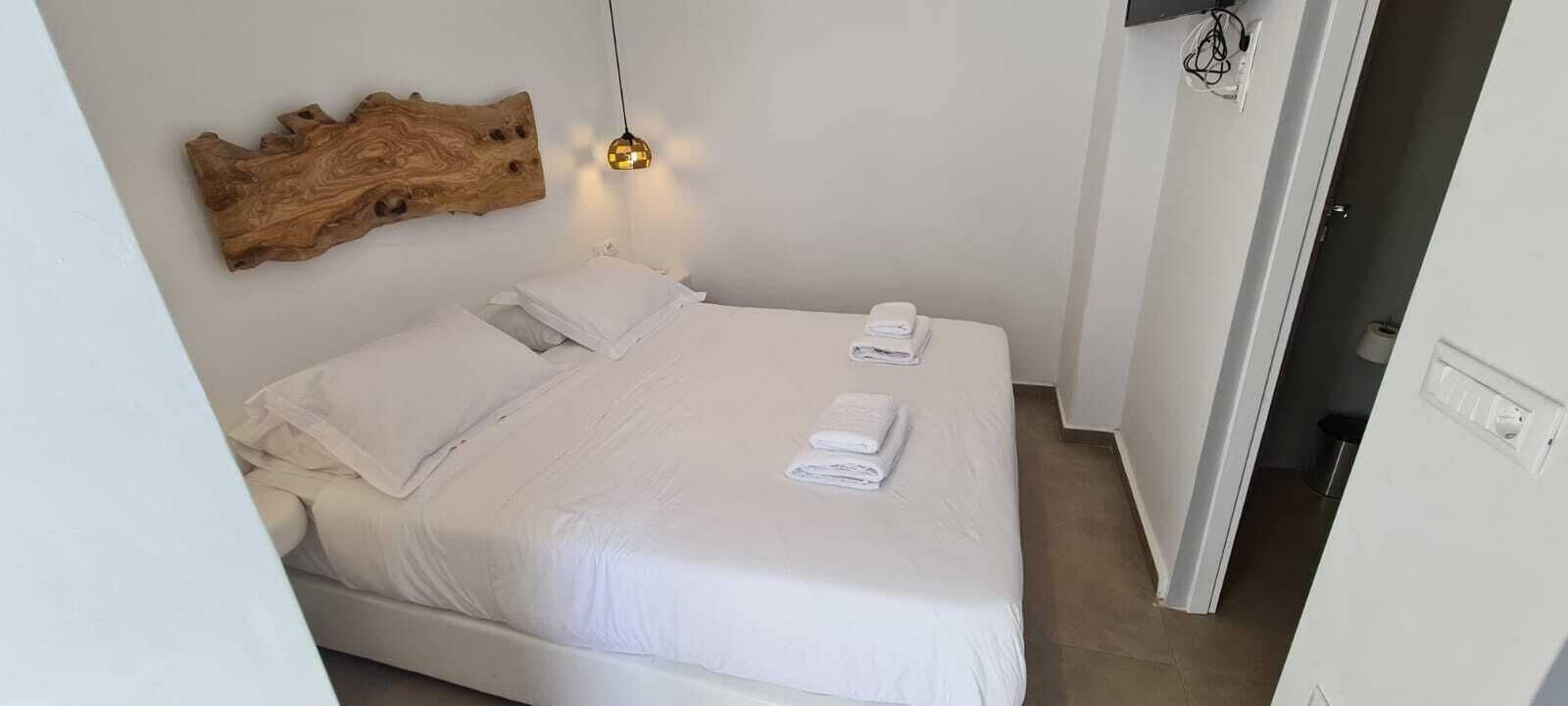 A neatly made bed with white linens and two folded towels, a wooden wall art piece, and a hanging light fixture.
