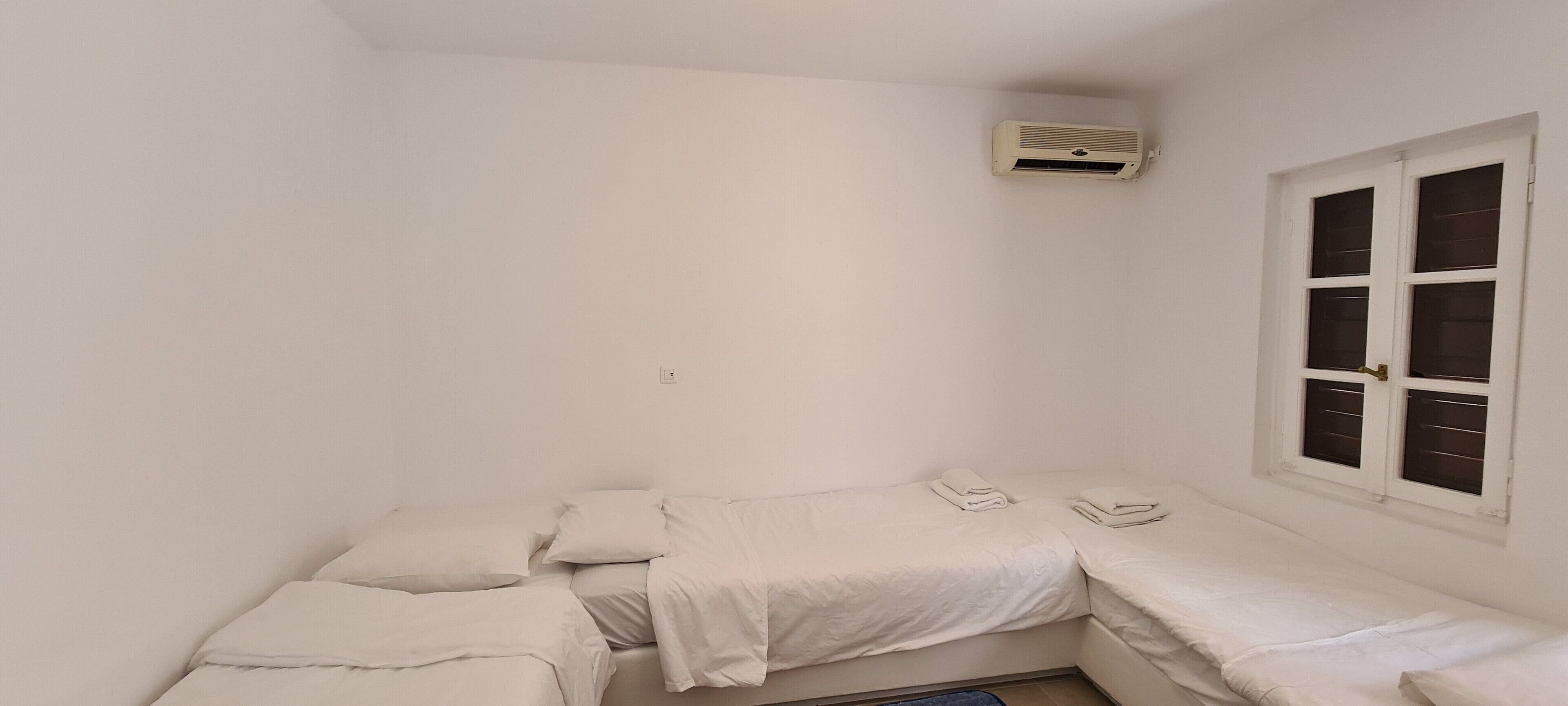 A small, white room with a single bed, a window with blinds, and an air conditioning unit on the wall.