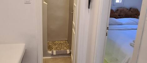Un couloir étroit, avec un lit au fond, un téléphone fixé au mur et une fenêtre avec des stores.