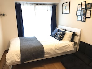 A neatly made bed with a gray blanket and two decorative pillows, a dark blue sofa, and a window with curtains.