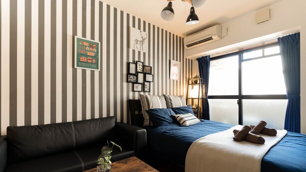 A room with a striped wall, a bed with a blue bedspread, a black leather sofa, a wooden table with a remote control, and a window with curtains. - Apartment Y Legendoal Nipponbashi (Osaka)