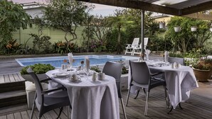 Un área de comedor junto a la piscina con mesas preparadas para dos, rodeada de abundante vegetación y una piscina.