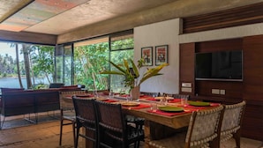 Un comedor con una mesa grande dispuesta para cuatro personas, rodeada de sillas de mimbre, una planta en maceta y un televisor montado en la pared.
