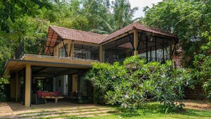 Una casa de dos pisos con un balcón, rodeada de abundante vegetación y palmeras.