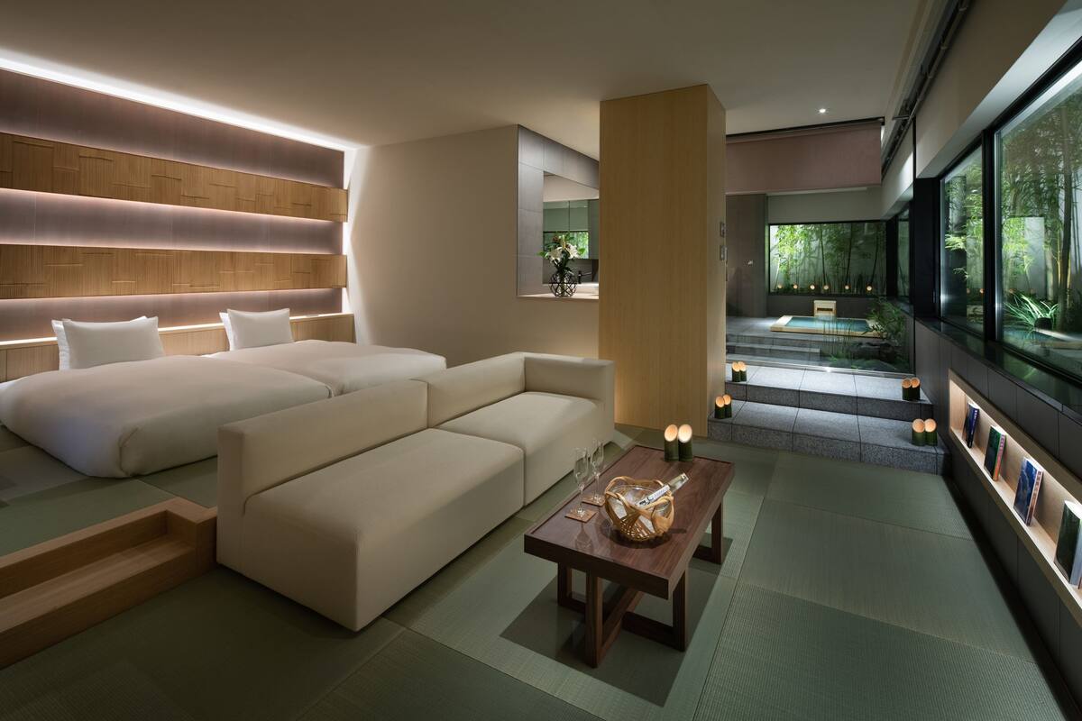Une chambre d’hôtel moderne avec un grand canapé, une petite table sur laquelle se trouvent un panier et des verres, une tête de lit en bois et une vue sur un jardin, visible à travers de grandes fenêtres.
