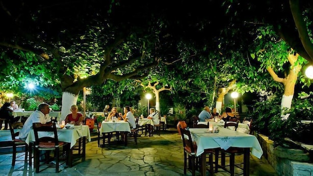 An outdoor dining area with tables and chairs under trees, surrounded by greenery. - Julia Apartments (Rethymno)
