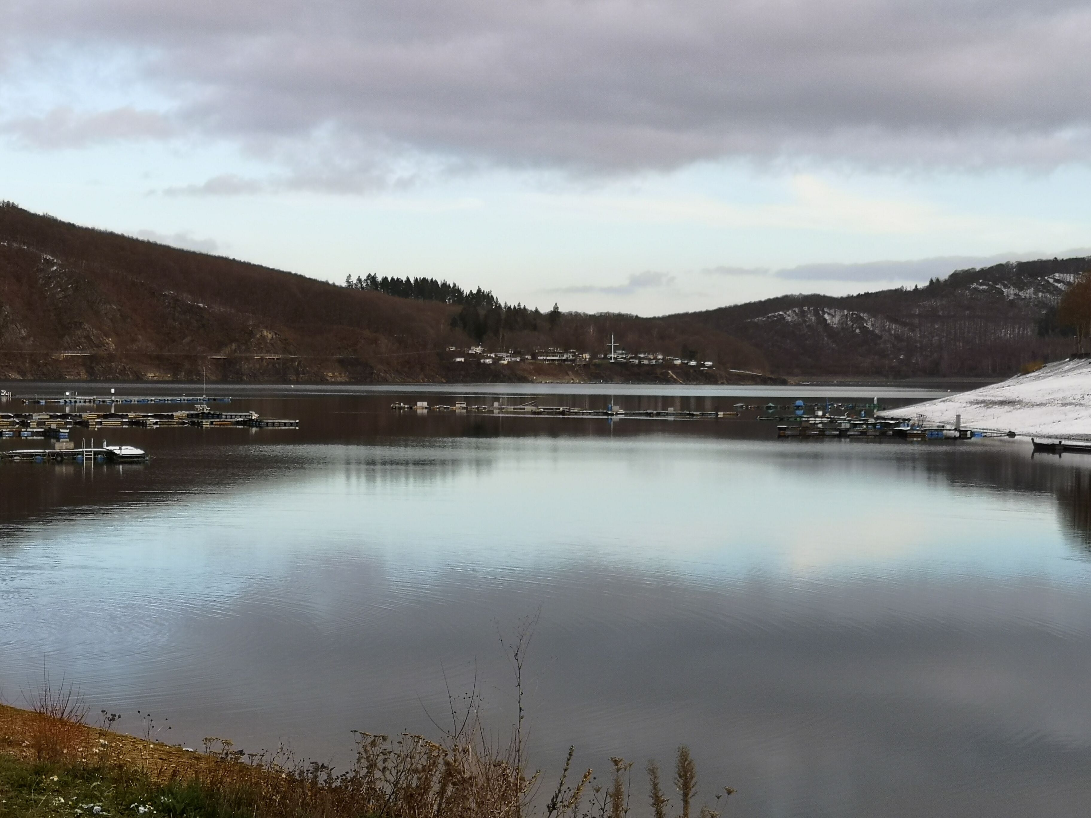 A calm lake with docks and boats, surrounded by hills and a small settlement.