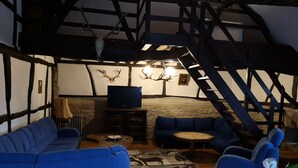 A rustic room with exposed wooden beams, a stone fireplace, a television on a wooden stand, blue upholstered seating, and a staircase. - Ferienhof Schmickerath (Simmerath)