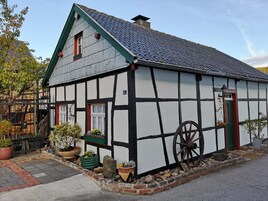 Una casa tradicional con entramado de madera, techo empinado, una gran rueda de madera y un pequeño jardín con plantas en macetas.