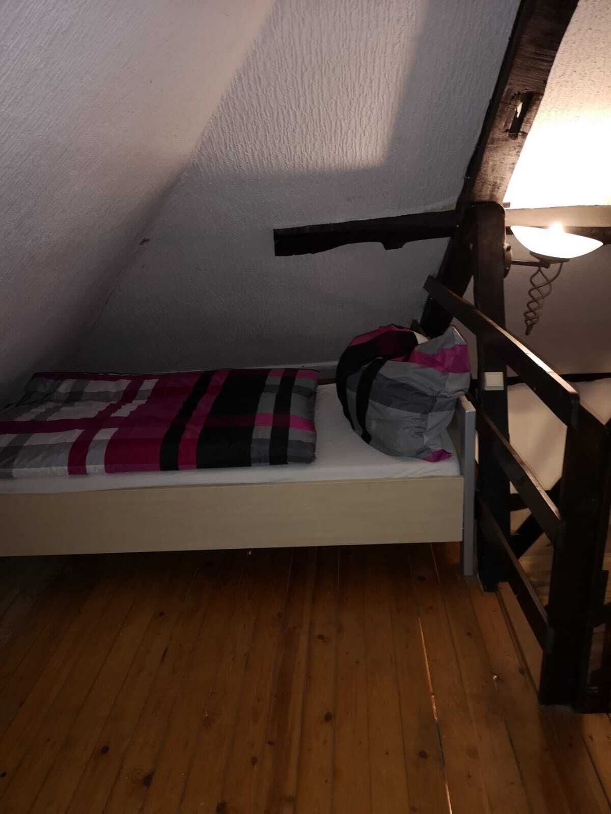 A small, narrow room with a wooden floor, a single bed with a checkered blanket, and a nightstand with a lamp.