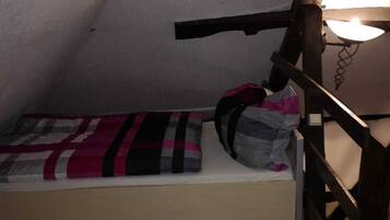 A small, narrow room with a wooden floor, a single bed with a checkered blanket, and a nightstand with a lamp.