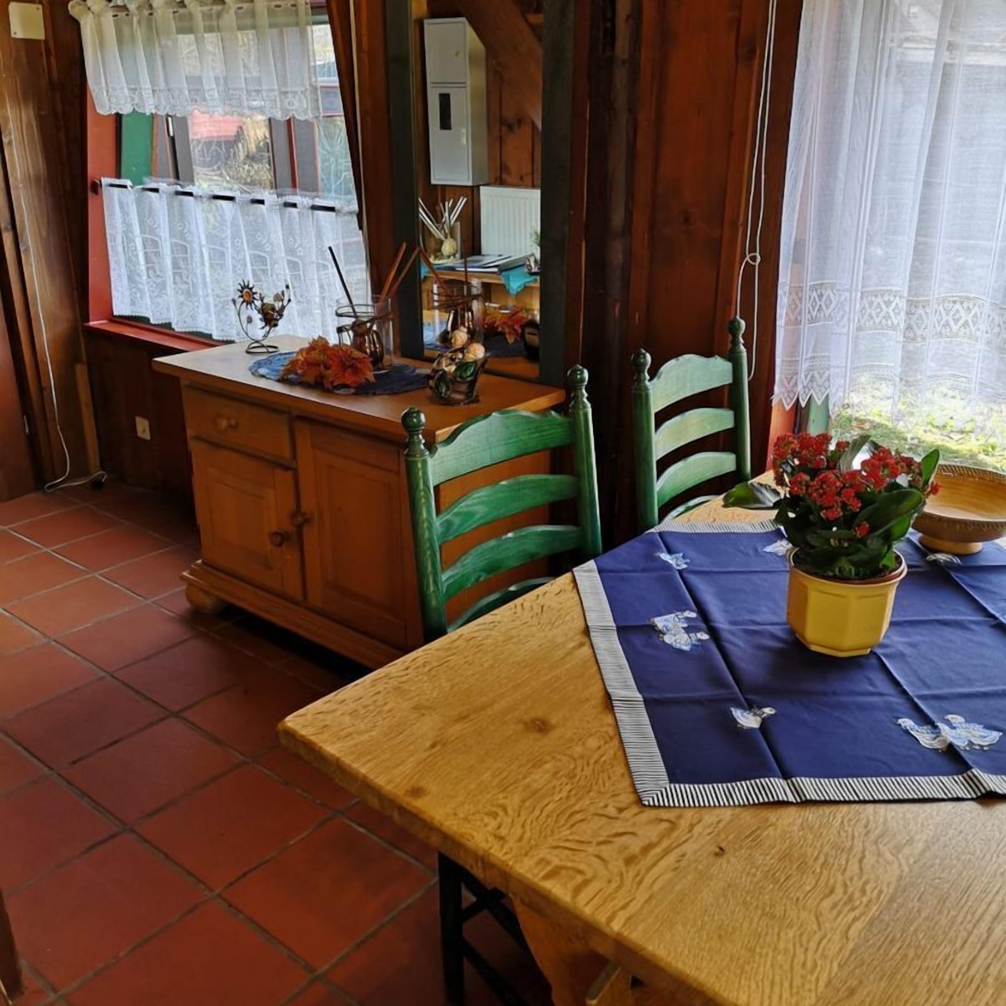 A dining area with a wooden table and chairs, a window with lace curtains, and a tiled floor.