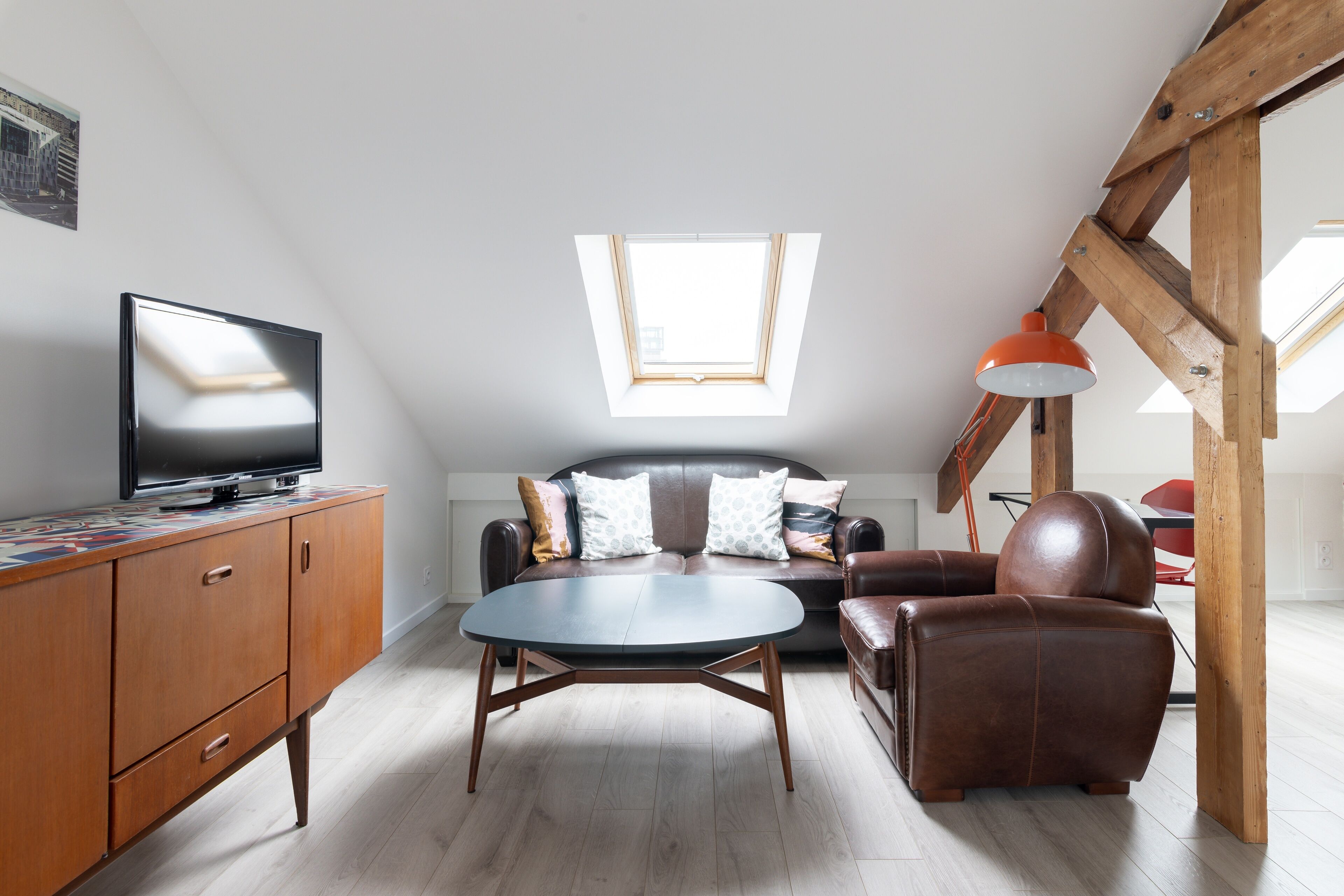 A modern loft apartment with a wooden ceiling, a flat-screen TV on a wooden cabinet, a brown leather sofa, a round coffee table, and a skylight.