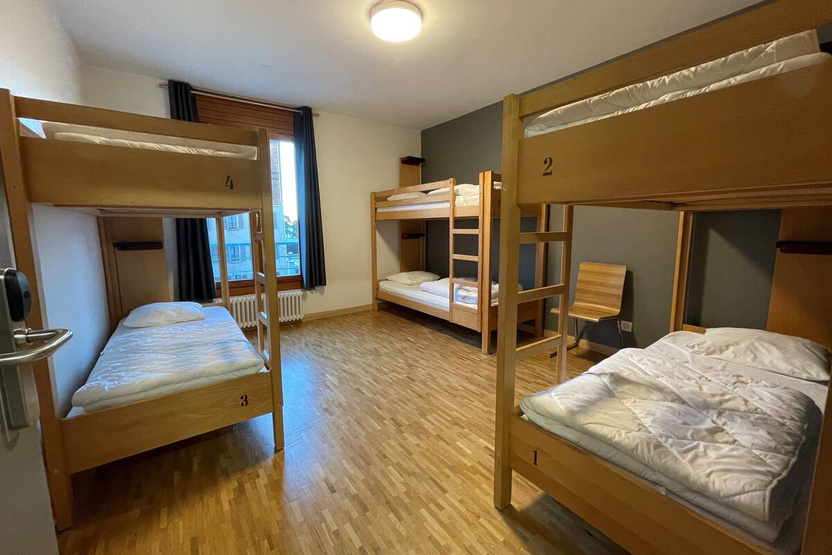 Asuntolan huone, jossa on kerrossängyt, puulattia, patteri, ikkuna verhoilla ja ovi, jossa on kahva.