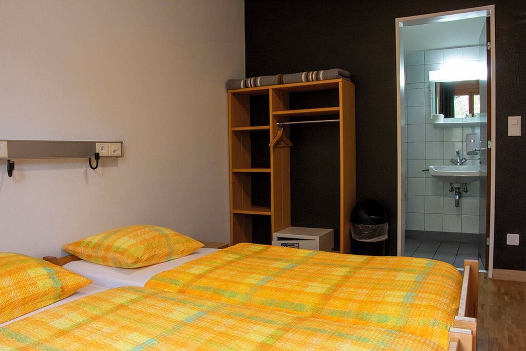 Un dormitorio con cama, armario, mesita de noche pequeña y baño visible a través de una puerta abierta.