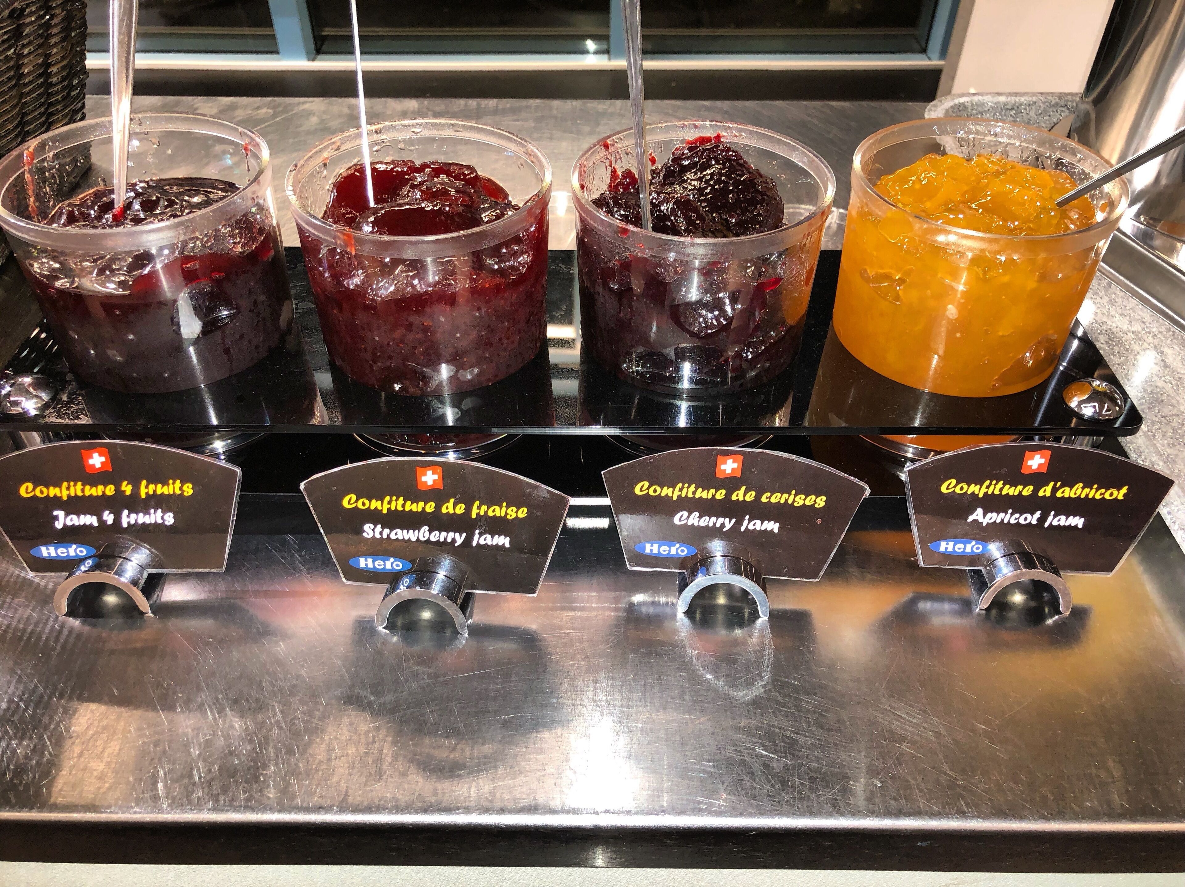 Una exhibición de alimentos con cuatro tipos distintos de mermeladas en vasos transparentes, cada una identificada con el tipo de mermelada y la marca 'Hero'.
