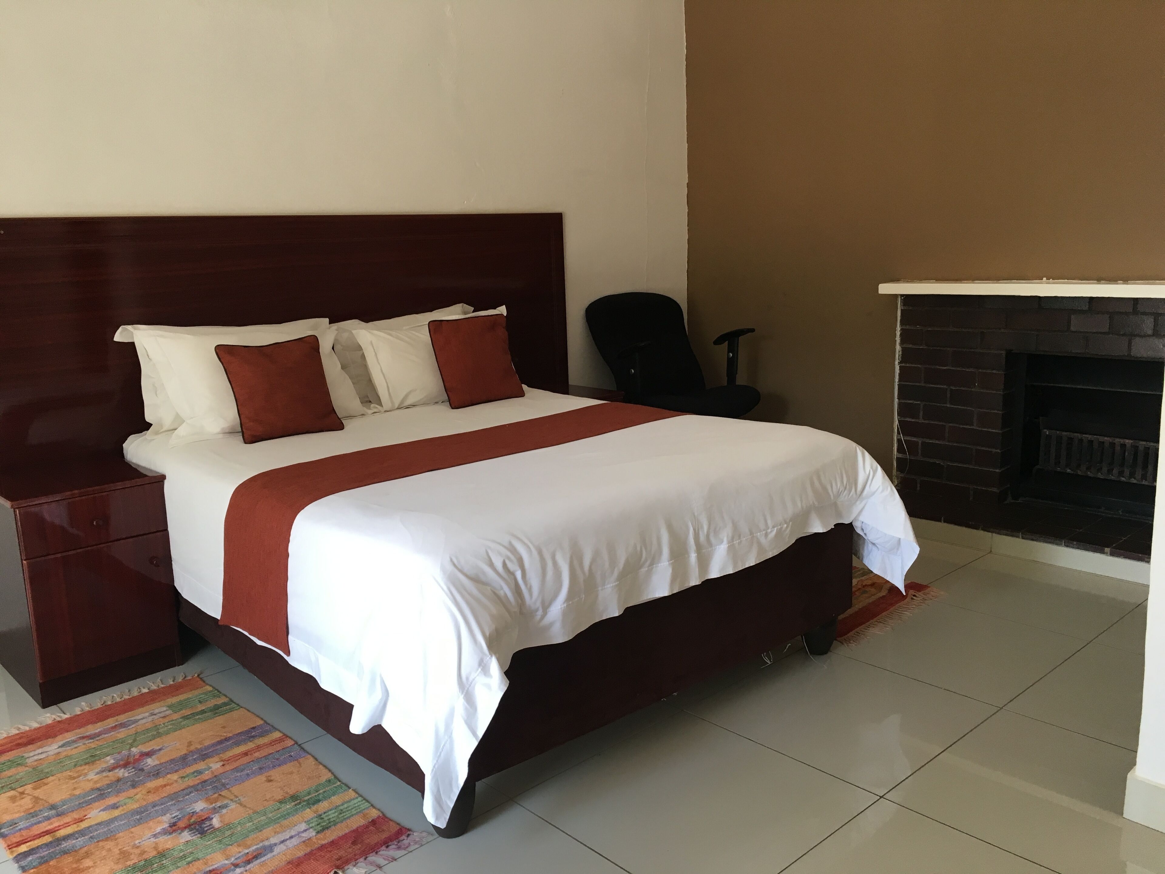 Una cama bien tendida con ropa de cama blanca y roja, un cabecero de madera, una silla de oficina negra y un suelo de baldosas.