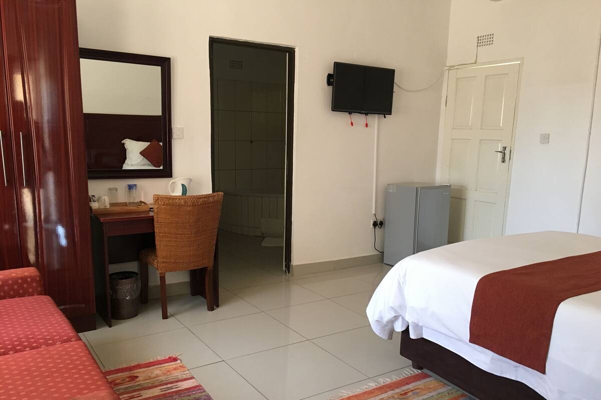 A hotel room with a bed, a desk with a chair, a television mounted on the wall, and a door leading to another room.