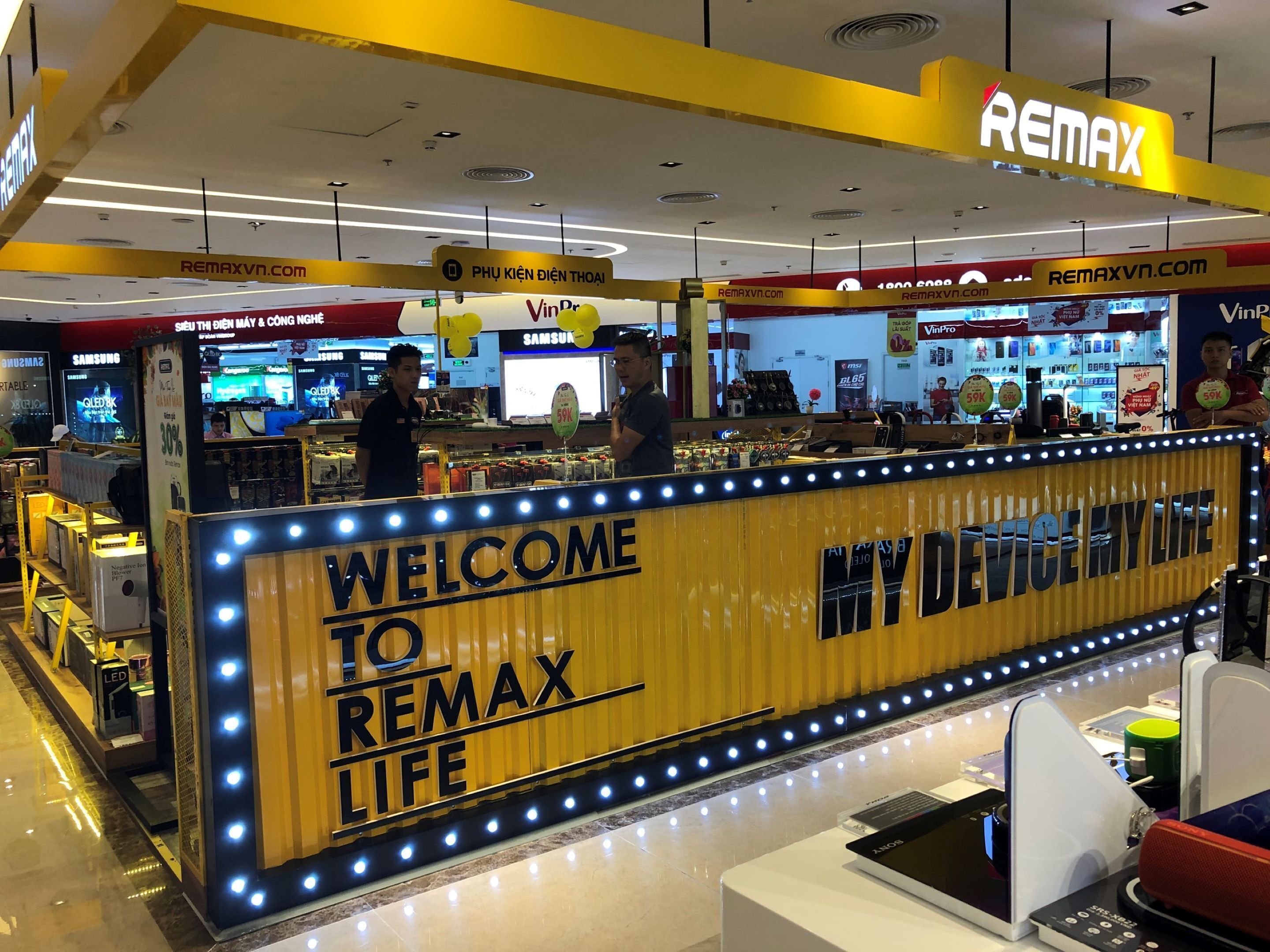 A store interior with a prominent 'Welcome to Remax Life' sign, various electronic devices on display, and a Samsung store in the background.
