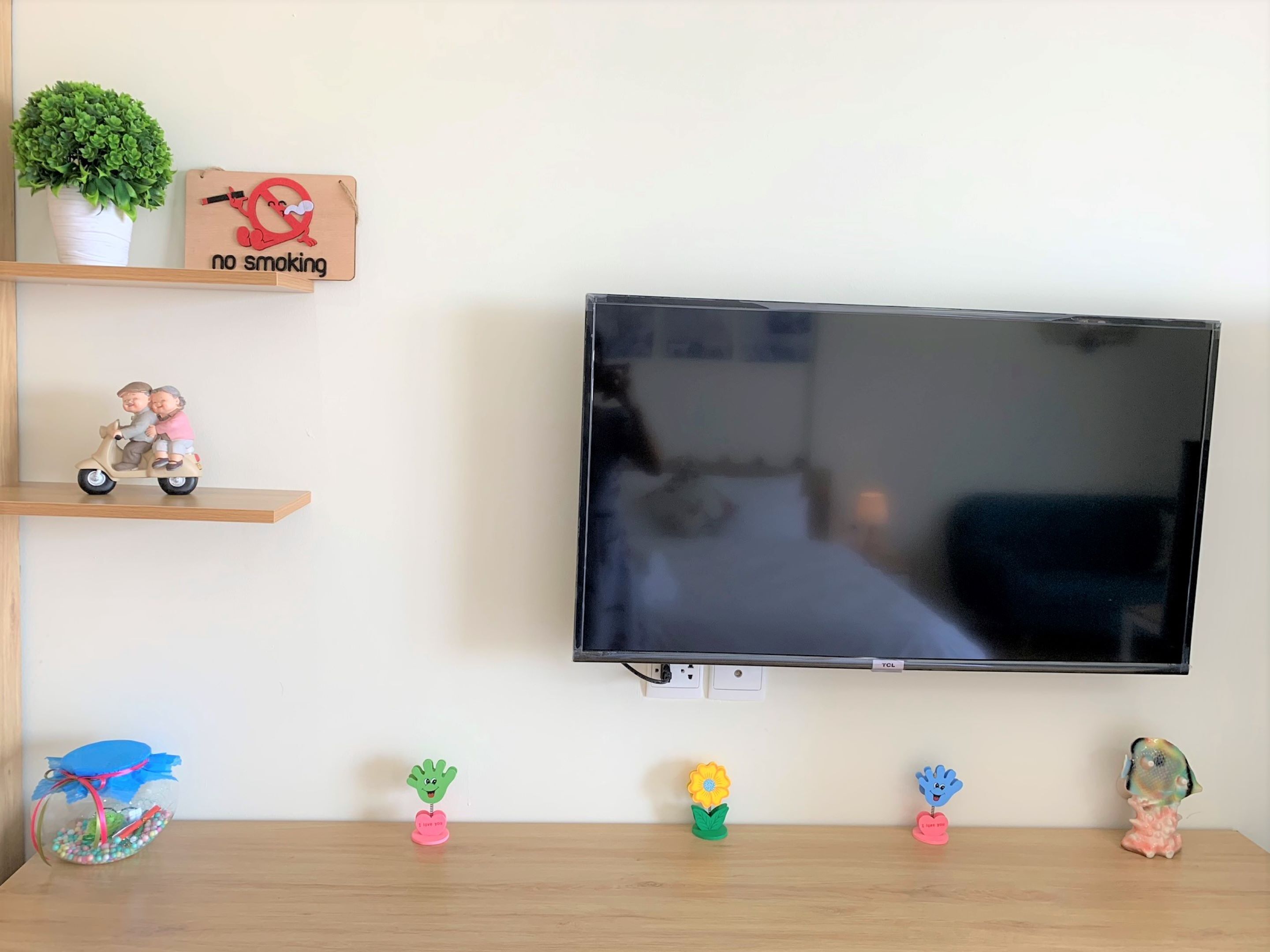A flat-screen TV mounted on a wall, a small potted plant, a 'no smoking' sign, a figurine of two people on a scooter, and decorative items on a shelf below.