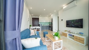A modern living room with a sofa, coffee table, and TV. There is a kitchen area with cabinets and a refrigerator.