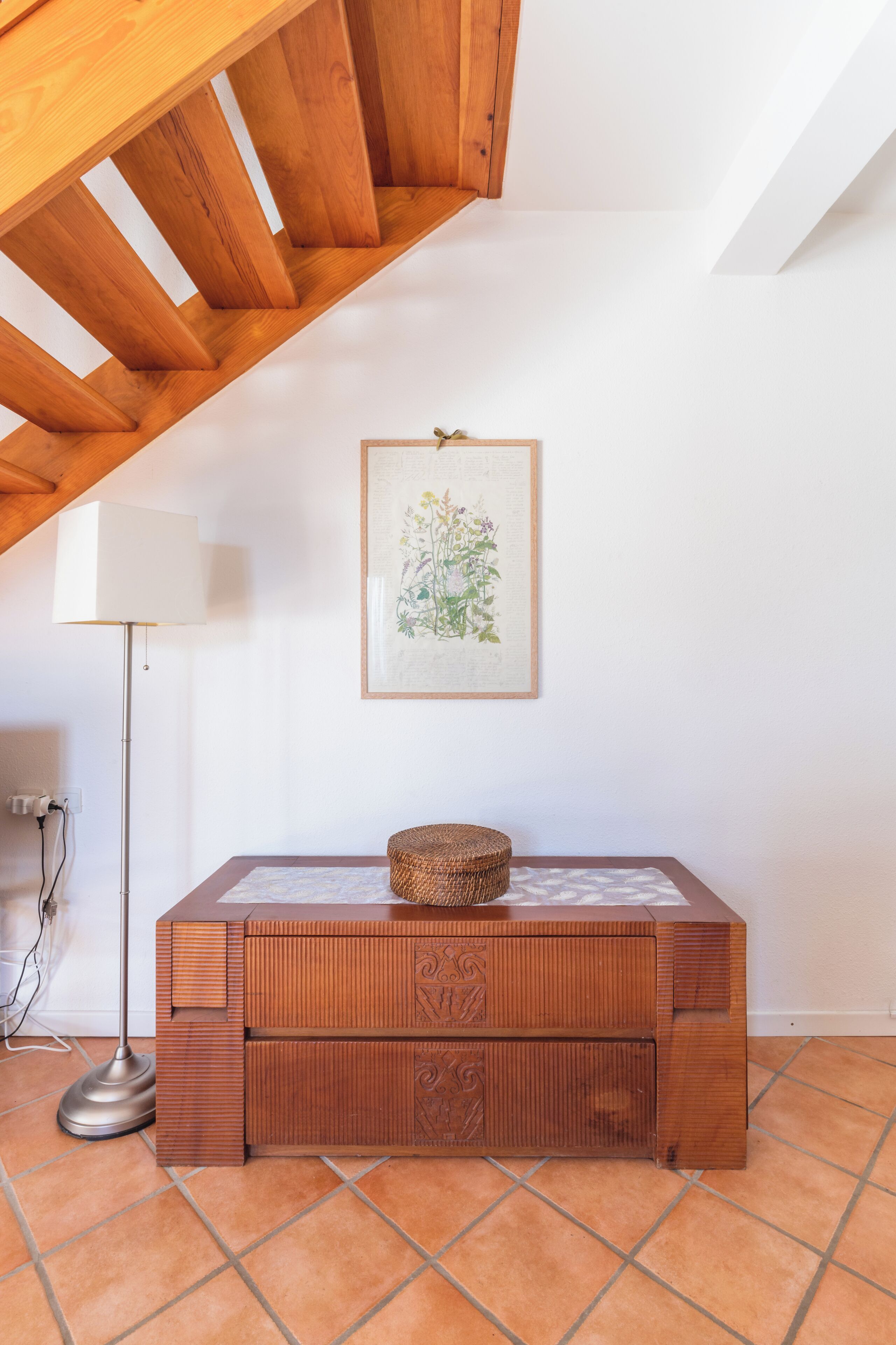 A room with a wooden bench, a framed botanical illustration, a floor lamp, and a tiled floor.