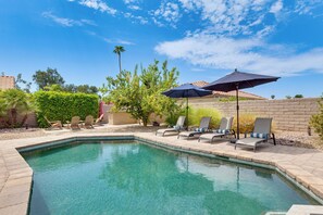 A swimming pool with clear water, surrounded by lounge chairs and umbrellas, and a brick wall in the background.