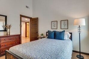A bedroom with a bed, dresser, bedside table, lamp, and framed botanical prints on the wall.