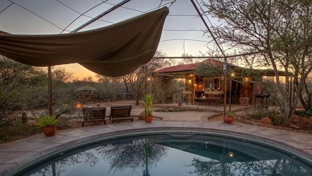 Zona de piscina con una pérgola, mobiliario exterior y un edificio de techo rojo al fondo.