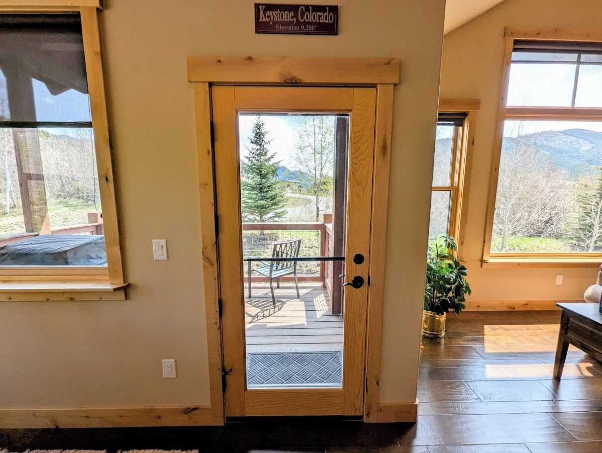 A wooden door with a glass panel leading to an outdoor deck with a bench. A sign above the door reads "Keystone, Colorado Elevation 9,280'".
