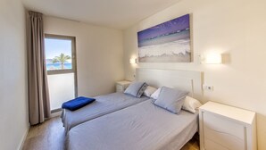 A bedroom with a large bed, two bedside tables, a window with curtains, and a painting of a beach scene. - APARTMENT IN CALA MILLOR, Modern, DIRECTLY ON THE BEACH, SEAVIEW, Wifi Incl, (Cala Millor)