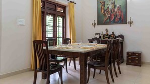 A dining area with a wooden table set for four, flanked by chairs, with a window and curtains on one side and a wall-mounted painting above the table. - Crystal Homestay by Vista Rooms (Virajpet)