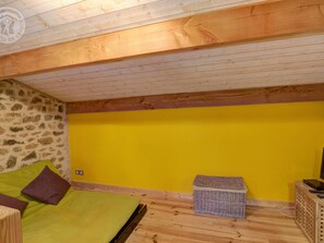 A room with a stone wall, a yellow wall, a wooden ceiling, a bed with green and brown bedding, a small wicker basket, and a wooden cabinet with a remote control.
