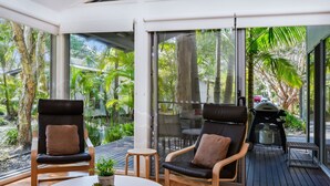 A covered outdoor seating area with two chairs and a small table, surrounded by glass doors and windows offering views of tropical vegetation. - Mobys Beach House # 11 (Boomerang Beach)