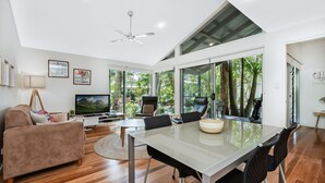 A modern living room with a flat-screen TV, a sofa, a dining table with chairs, and a ceiling fan. - Mobys Beach House # 11 (Boomerang Beach)