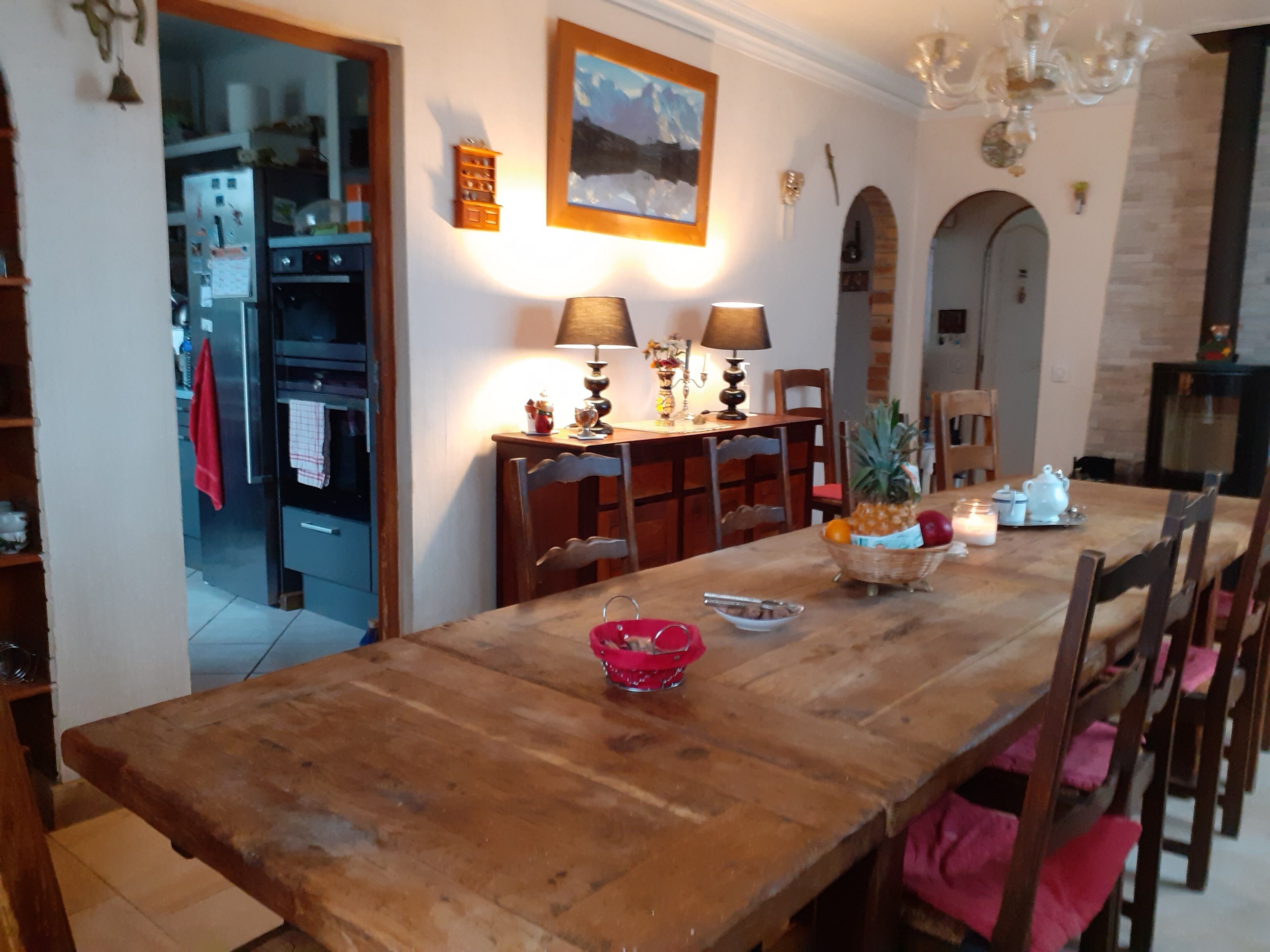 A dining area with a wooden table, chairs, and a chandelier. There is a kitchen in the background with a refrigerator and cabinets.