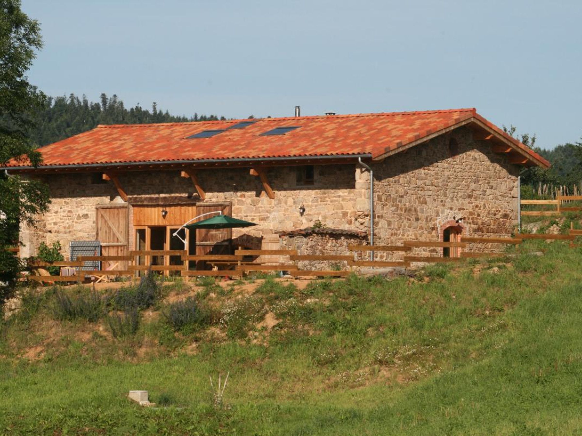 Un edificio rústico de piedra con techo de tejas rojas, un porche de madera y un patio cercado.