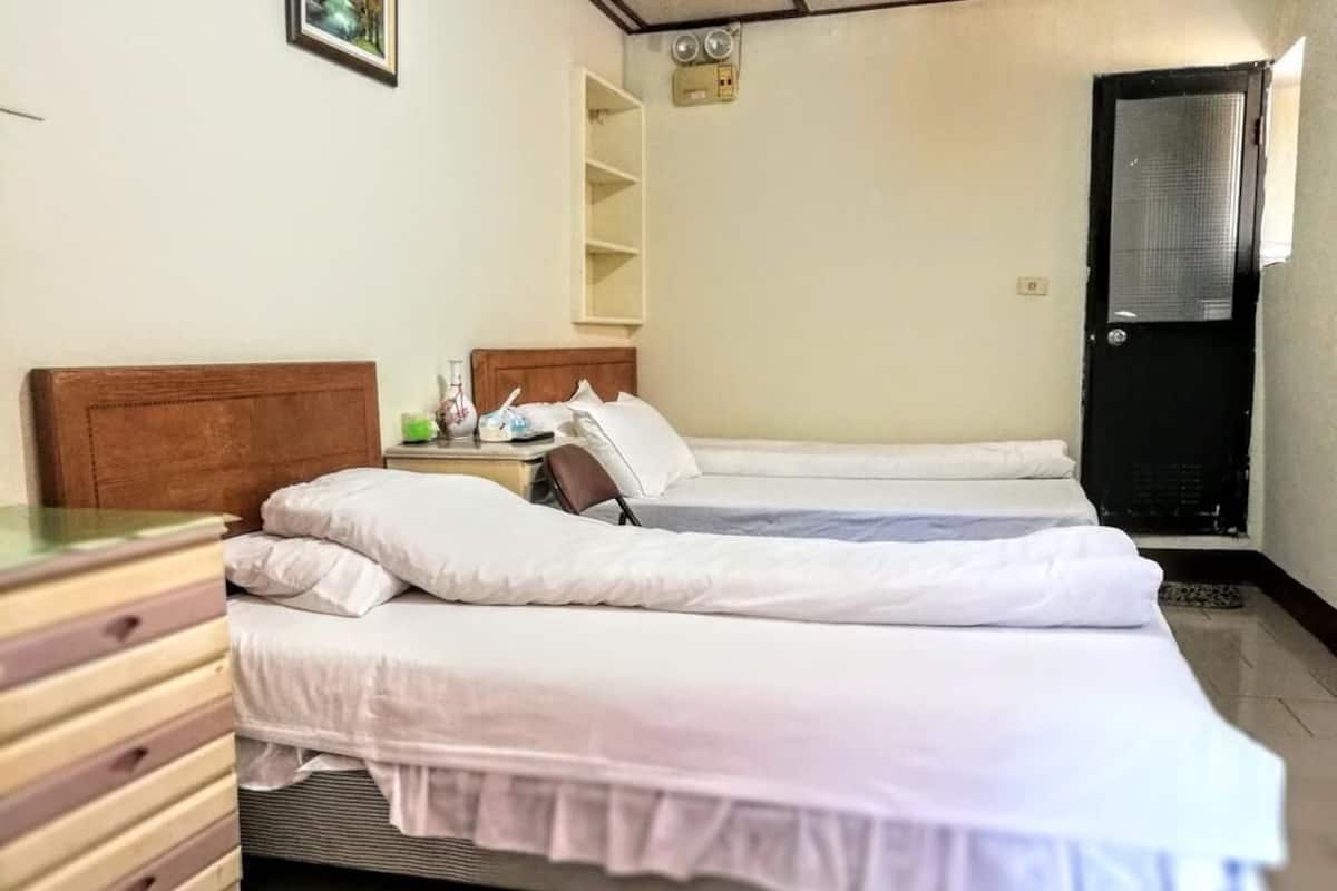 Habitación de hotel con dos camas individuales, un tocador de madera, una mesita de noche, una estantería y una puerta.