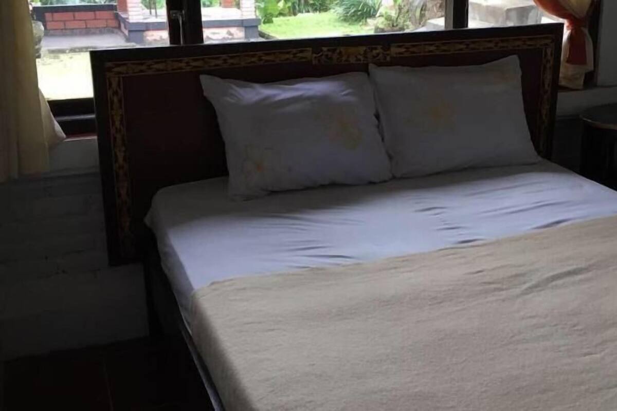A bed with a headboard, a window with a view of greenery, and a small round table.