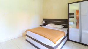 A neatly made bed with a white and orange blanket, a bedside table with a lamp, and a large mirror on the wall. - Bantan Guest House (Tibubeneng)