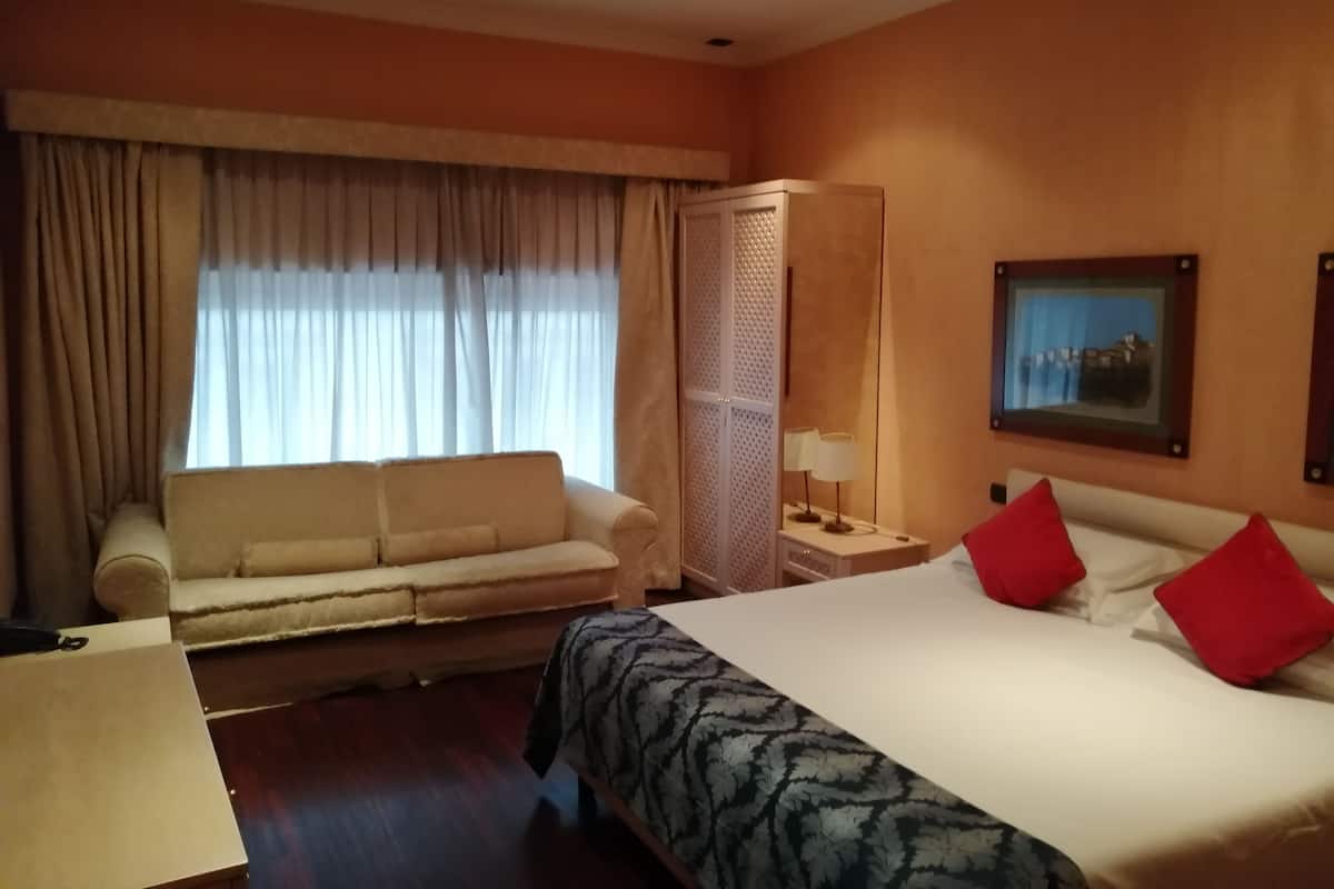 A hotel room with a bed, a sofa, a nightstand with a lamp, a wardrobe, and a painting on the wall.