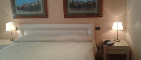 A hotel room with a bed, two bedside lamps, a nightstand with a phone, and two framed paintings on the wall.