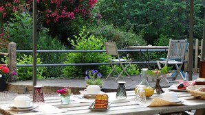 An outdoor dining area with a wooden table set for breakfast, surrounded by lush greenery and a metal railing. - Moulin de la Papeterie Chambres d'hôtes (La Creche)