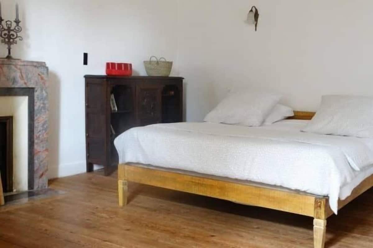 A bedroom with a wooden bed, white bedding, a nightstand, a wooden wardrobe, and a small table with a red bowl and a woven basket.