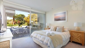 A bedroom with a large bed, a bedside table with a lamp, a glass door leading to a patio with outdoor furniture, and a painting on the wall.